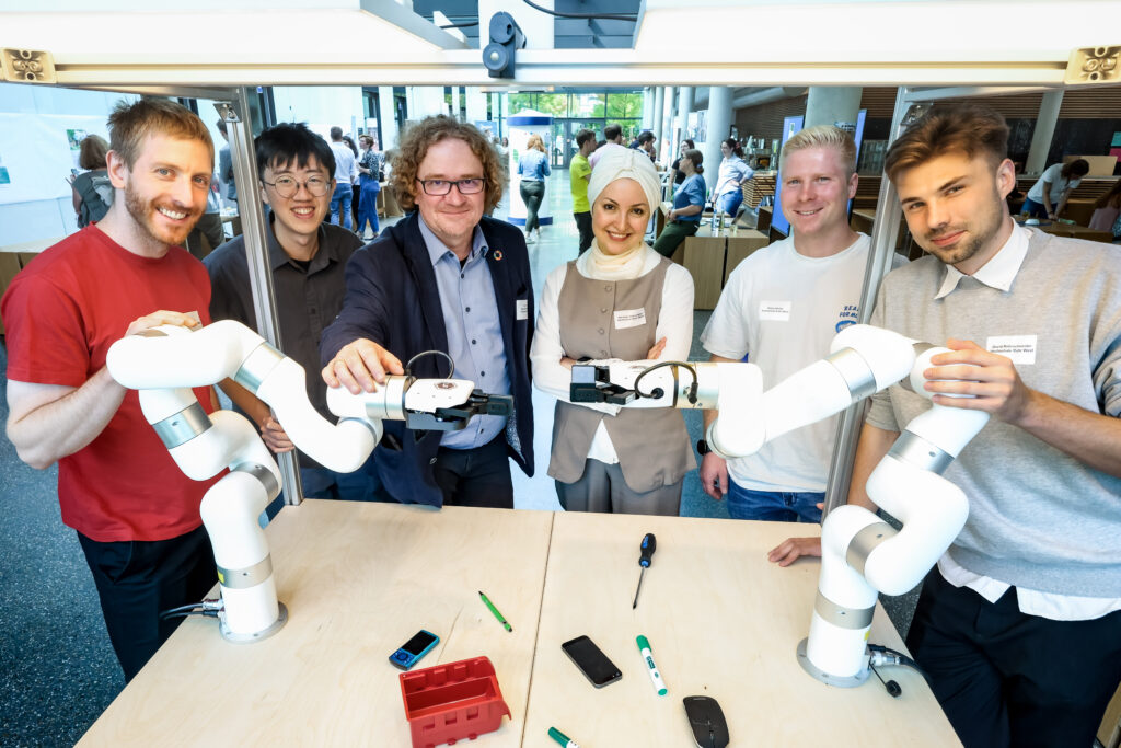 Das Bild zeigt sechs Personen vor zwei weißen Roboterarmen. Es handelt sich um das Team KI des Transferhubs von Prof. Uwe Handmann.