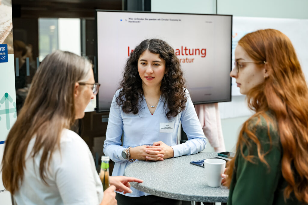 Drei Frauen stehen an einem Stehtisch. Es ist der Infostand zum Handwerk und Digital Circular Economy. Dahinter ein Bildschirm auf dem Instandhaltung und Reparatur steht.