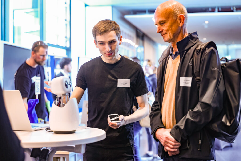 Zwei Männer einer jünger, einer älter stehen neben einem Stehtisch. Auf diesem Stehtisch steht ein kleiner Roboter. Dr jüngere Mann links erklärt etwas zu dem Roboter. Beide sehen freundlich und interessiert aus.