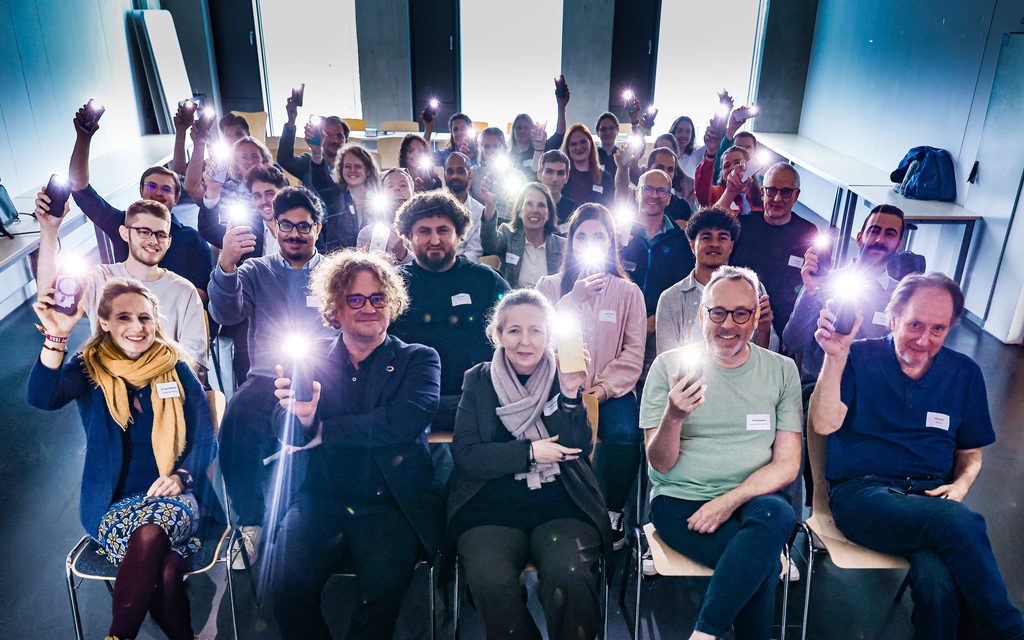 Das Publikum der Abschlusssession des Barcamps 2026 hält in einem dunklen Raum Smartphones mit Taschenlampe hoch. Alle lachen nach vorne.