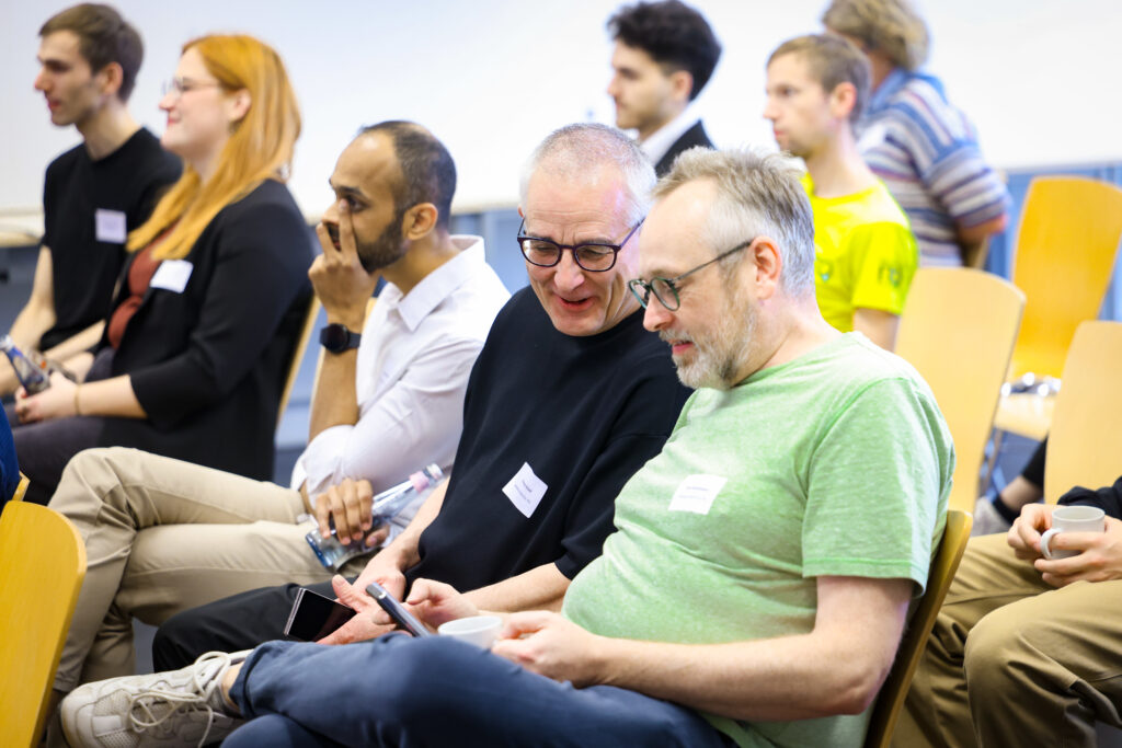 Das Publikum des Barcamps stimmt ab. Im Vordergrund zwei Männer die lachend auf einen Handybildschirm blicken und voten.