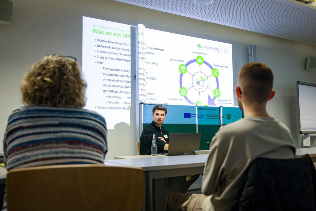 Ein junger Mann sitzt vorne in einem Seminarraum. Im Hintergrund steht was ist ein DPP und Foliennotizen dazu mit einer Grafik. Er spricht zu zwei Menschen, die ihn anschauen. Eine ältere Frau und ein junger Mann, diese sieht man nur von hinten.