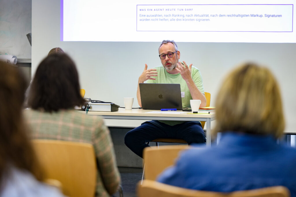Ein Mann in einem hellen grünen Tshirt sitzt an einem Tisch. Hinter ihm ist zu lesen: "Was ein Agent heute tun darf? Eine auswählen, nach Ranking, nach Aktualität, nach dem reichhaltigsten Markup. Signaturen würden nicht helfen, alle drei konnten signieren."
Er spricht mit Teilnehmerinnen, sie sitzen mit dem Rücken zur Kamera.