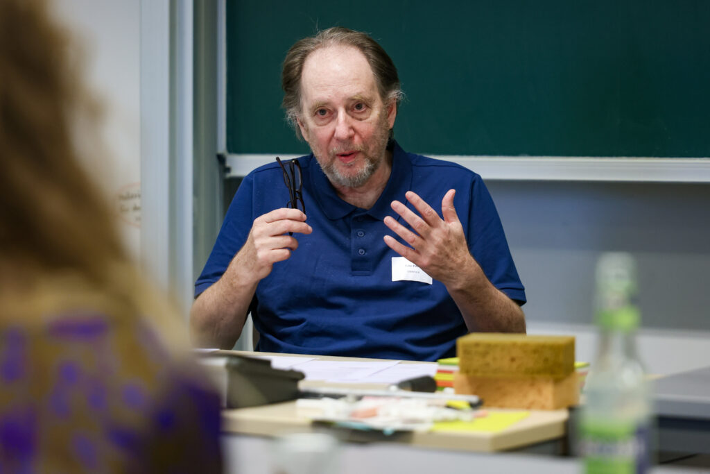 Peter Flesch beim Barcamp 2026. er spricht zur Audience. Hinter ihm eine Tafel. Er hält seine Brille in der Hand.
