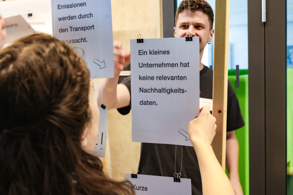 Stand Neue Effizienz: Es werden Mythen gezeigt z.B. "Ein kleines Unternehmen hat keine relevanten Nachhaltigkeitsdaten", auf der Rückseite steht die Auflösung, das wird klar durch den Pfeil auf dem Papier. Eine Frau interagiert damit. Ein Mann lächelt im Hintergrund.