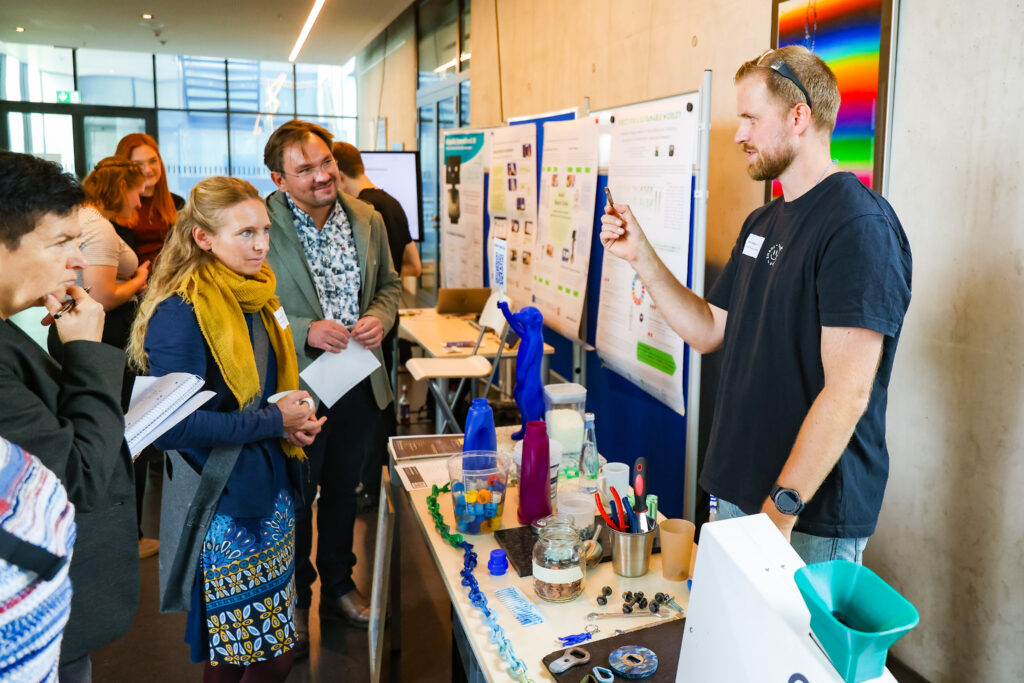 Zu sehen ist ein Mann der Plastikrecycling vor Ort einer Gruppe von Menschen vorstellt. Diese schauen gespannt zu. Eine Frau guckt sehr konzentriert, ein Mann lächelt. Im Hintergrund sind Stellwände zu sehen mit wissenschaftlichen Postern.
