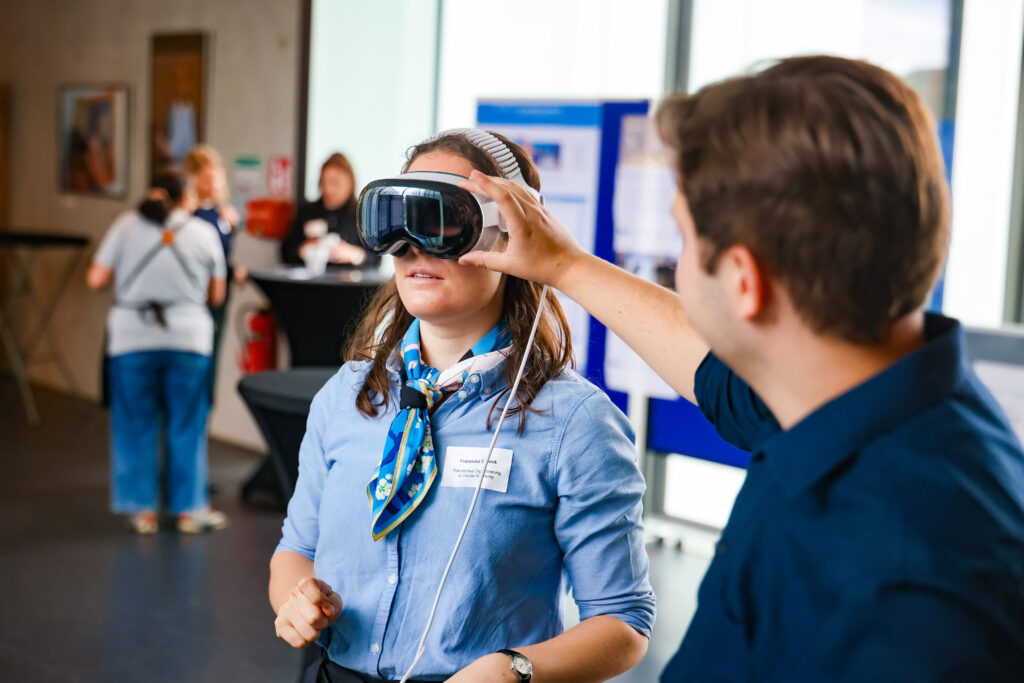 Eine Frau trägt eine VR Brille ein Mann hilft ihr diese anzupassen.